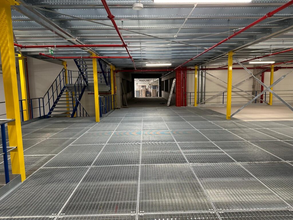 Entresolvloer op maat met stalen roosterplaten, gele kolommen en trap, montage in magazijn